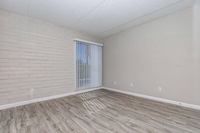 Modern room featuring elegant wood-look flooring, a stylish brick accent wall, and large window with vertical blinds, inviting ample natural light.