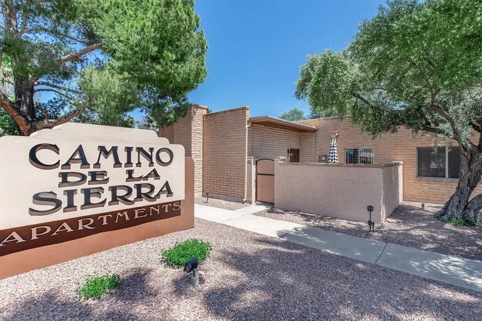 Charming entrance to Camino De La Sierra Apartments, framed by lush trees and a sunny brick facade.