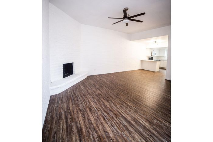 Chic living room with elegant dark wood flooring, cozy white brick fireplace, and modern ceiling fan, seamlessly leading to a bright, open kitchen.