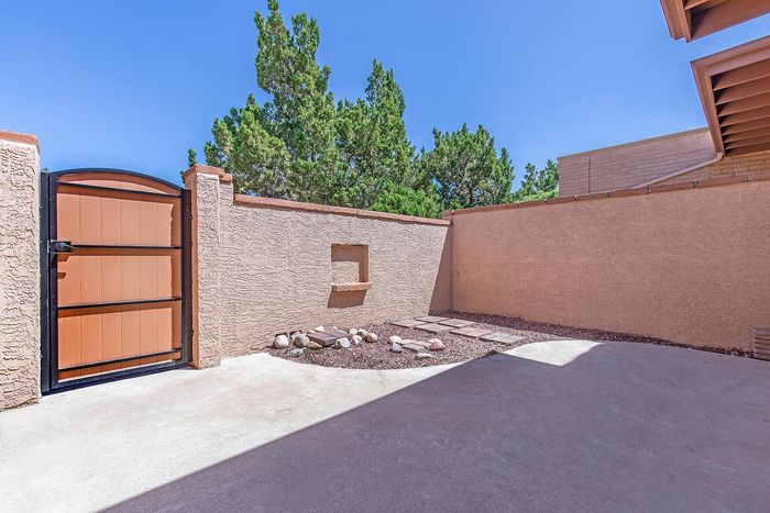 Charming outdoor patio with rustic wooden gate, bright stone walkway, and lush greenery under a clear blue sky.