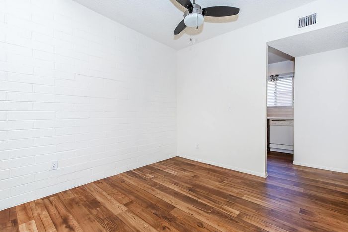 Bright, spacious room with elegant wood flooring and a modern ceiling fan, leading to a sunlit kitchen area.