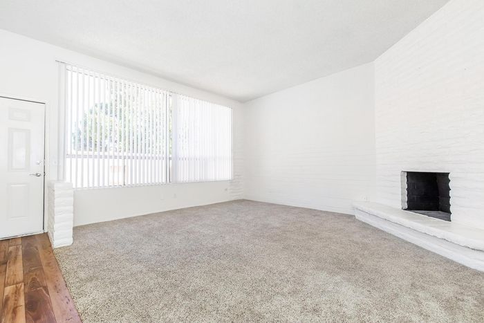 Bright living room with plush carpet, large windows with vertical blinds, and a modern white brick fireplace.