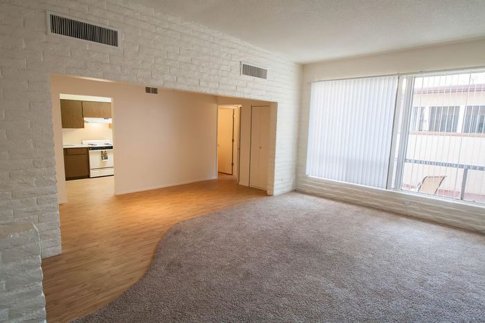 Bright and spacious living room with large windows, cozy carpet, and an open layout leading to the kitchen, perfect for modern living.
