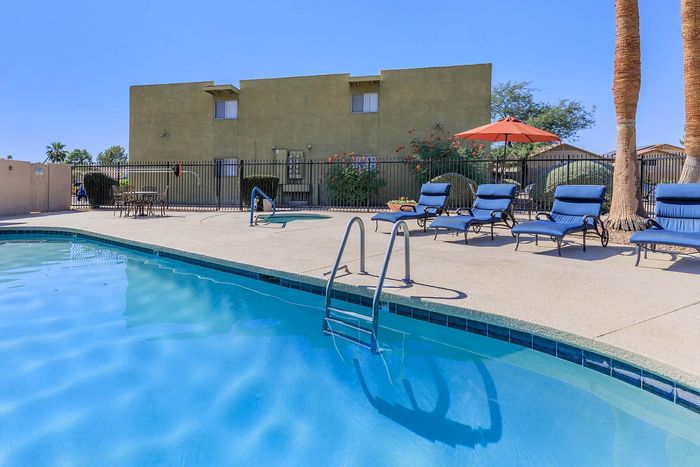 Inviting outdoor pool area with comfortable lounge chairs and a large umbrella, framed by palm trees and a well-maintained building in the background.