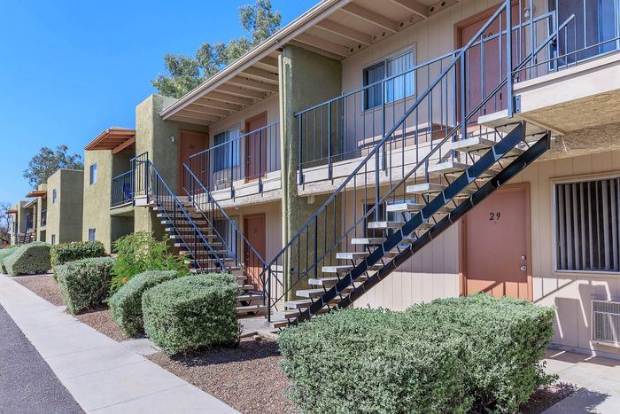 Charming apartment complex exterior with lush greenery, modern staircase access, and vibrant doors under clear blue skies.