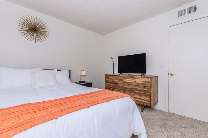 Elegant bedroom with a cozy bed adorned with a bright orange throw, stylish wooden dresser, and a sleek flat-screen TV, perfect for relaxing and unwinding.