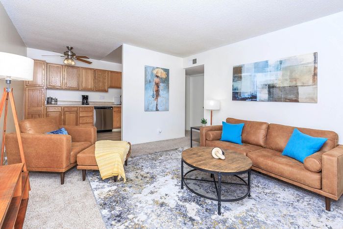 Inviting living room with cozy brown leather seating, vibrant blue cushions, and elegant wooden coffee table, opening to a sleek kitchen with modern cabinetry.