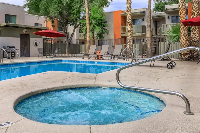 Inviting apartment complex pool area with refreshing hot tub, cozy lounge chairs, and vibrant umbrellas surrounded by lush palm trees.