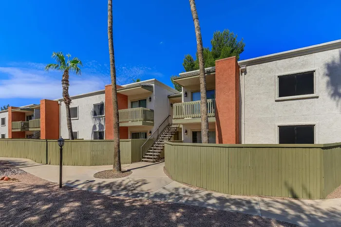 Charming modern apartment complex with vibrant orange accents, lush palm trees, and private balconies against a clear blue sky.