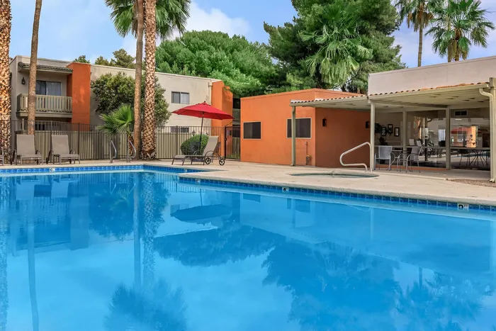 Vibrant apartment pool area showcasing crystal-clear water, palm trees, and comfortable lounge chairs with red umbrella.