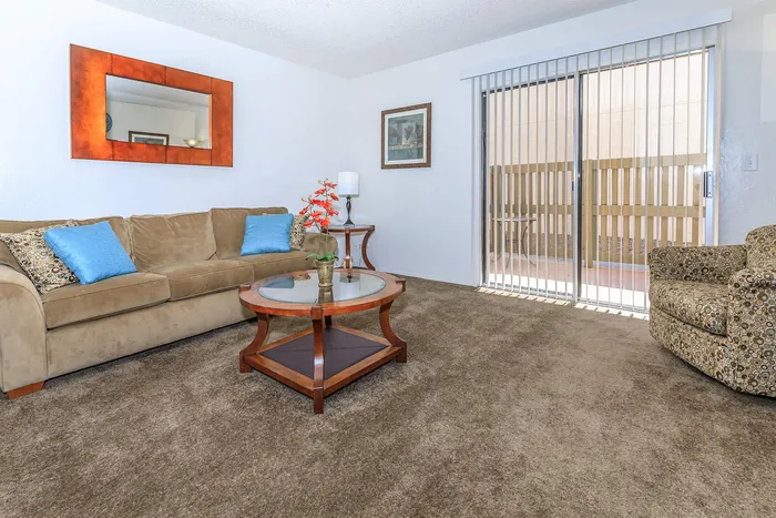 Inviting living room with plush carpet, cozy beige sofa adorned with bright blue cushions, elegant glass coffee table, and large window with vertical blinds for natural light.