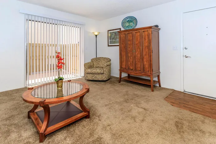 Inviting living room with cozy carpet, elegant glass-top coffee table, plush armchair, and rich wooden cabinet, bathed in natural light from large sliding doors.