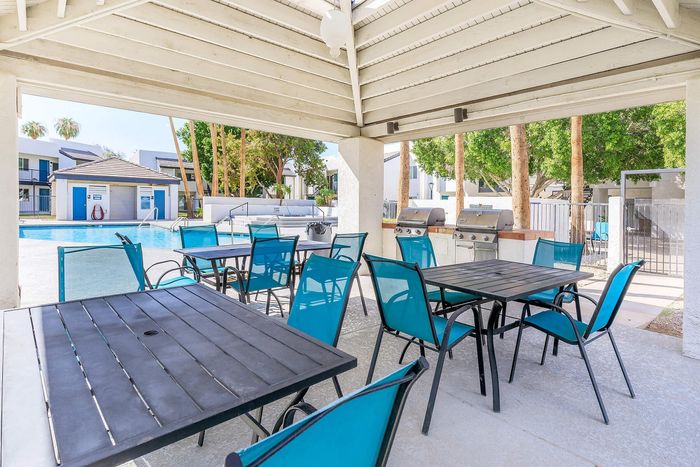 Charming outdoor patio with vibrant blue chairs, shaded under a white pergola, overlooking a sparkling community pool with barbecue grills nearby.