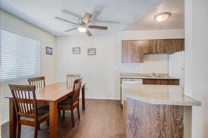 Charming dining area with a wooden table set beneath a ceiling fan, adjacent to a modern kitchen featuring sleek wooden cabinets and bright countertops.