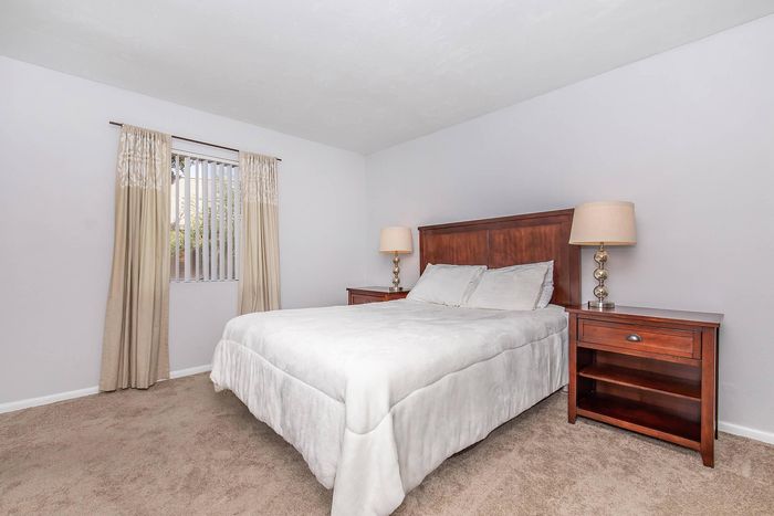 Bright and inviting bedroom with plush carpeting, elegant wooden furniture, and soft natural light filtering through large window curtains.