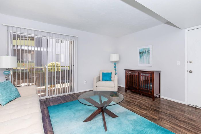 Bright and modern living room featuring sleek hardwood floors, a glass coffee table, and abundant natural light through vertical blinds.