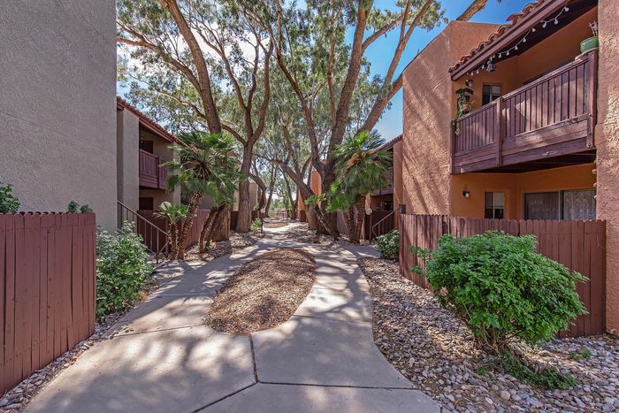 Charming apartment complex walkway shaded by lush trees and adorned with vibrant greenery, leading to cozy balconies.