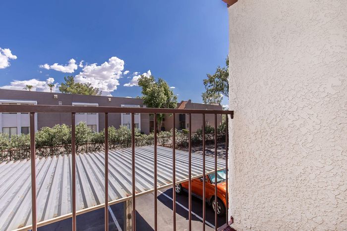 Bright patio view from the apartment, overlooking a covered parking area and lush greenery, under a vibrant blue sky.