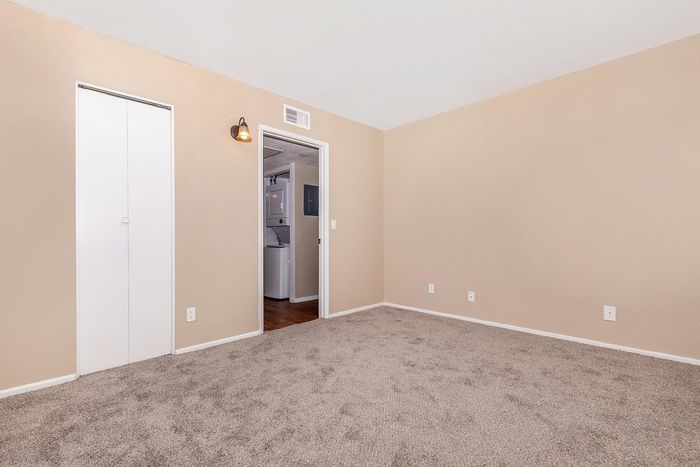 Bright and spacious bedroom with plush carpet and warm beige walls, featuring a closet and a view of a modern laundry area through the doorway.