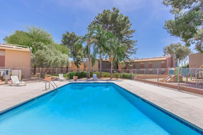 Inviting apartment complex pool area with vibrant blue water, surrounded by lush greenery and comfortable lounge chairs, perfect for relaxation under the sunny sky.