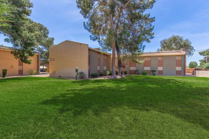 Charming apartment building with a well-maintained grassy lawn, surrounded by tall trees under a clear blue sky.