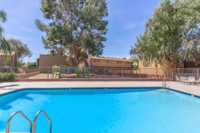 Inviting apartment pool area with crystal-clear blue water, surrounded by lush trees and a fenced playground, perfect for family relaxation.