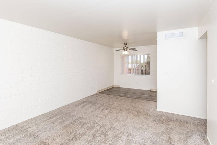 Bright and spacious living area with plush carpeting, featuring a stylish ceiling fan and large window with blinds for natural light.