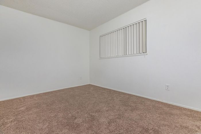 Bright and cozy carpeted room featuring soft neutral tones, large window with vertical blinds, and clean, minimalist design.