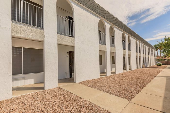 Elegant two-story apartment building with classic arches, spacious balconies, and a neat gravel landscape under a bright blue sky.
