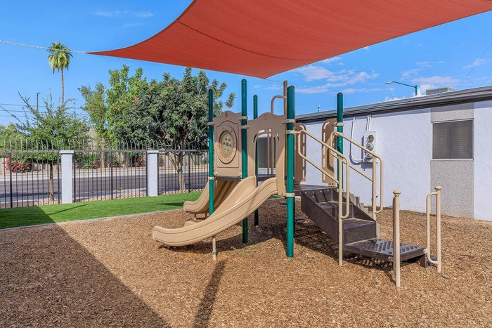 Charming playground with modern slides and shade sail, set against a backdrop of lush greenery and a sunny, blue sky.