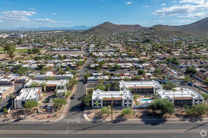 Aerial view of charming suburban neighborhood with modern white-roofed buildings, lush greenery, and picturesque mountain backdrop under a clear blue sky.
