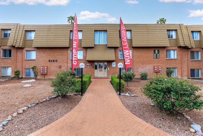 Inviting apartment complex entrance with modern brick fa&ccedil;ade, lush landscaping, and welcoming 'Now Leasing' banners.