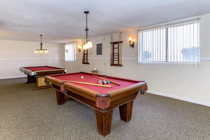 Inviting game room with two elegant billiards tables, warm lighting, and natural sunlight through large windows.