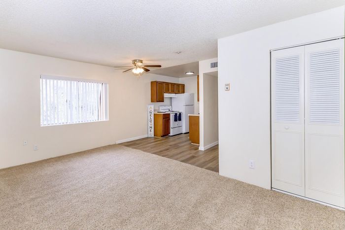 Bright open living space with plush carpeting, featuring a modern ceiling fan, large window for natural light, and cozy kitchen area with wood cabinets and white appliances.