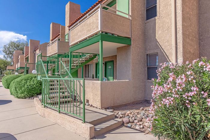 Charming desert-style apartment with vibrant green accents, outdoor staircase, and lush landscaping under a bright blue sky.