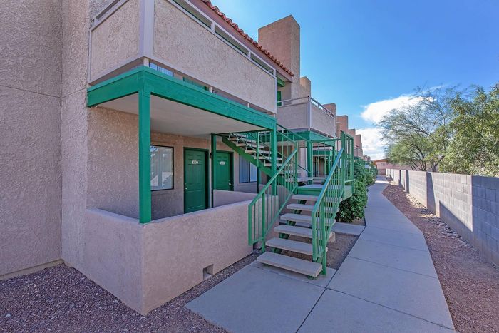 Charming apartment exterior with green accents, featuring an inviting stairway and lush landscaping under a bright blue sky.