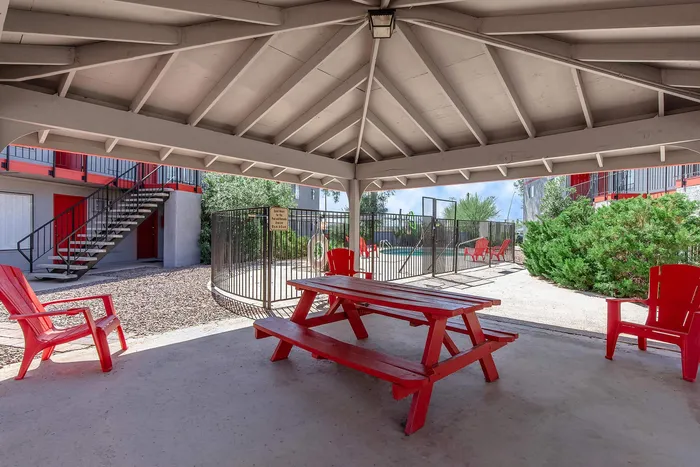 Charming outdoor communal area with vibrant red picnic seating under a cozy gazebo, overlooking a fenced pool and lush greenery.