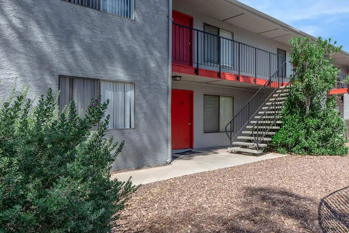 Charming apartment complex with vibrant red doors, lush greenery, and an inviting stairway under a clear blue sky.