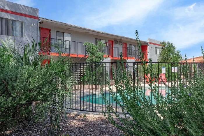 Charming apartment complex with a refreshing pool, vibrant red accents, and lush greenery under a bright blue sky.