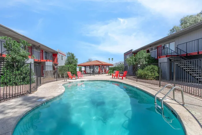 Inviting apartment complex featuring a sparkling swimming pool, vibrant red seating, and well-maintained landscaping under a clear blue sky.