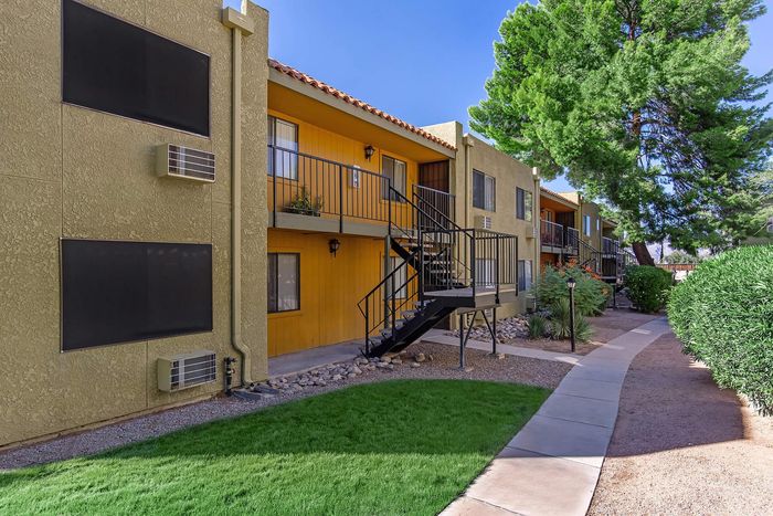 Charming apartment exterior with vibrant yellow accents, lush greenery, and inviting pathways nestled under tall trees.