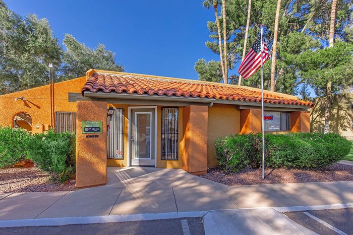 Charming Spanish-style apartment office with terracotta roof, surrounded by lush greenery and a welcoming American flag.