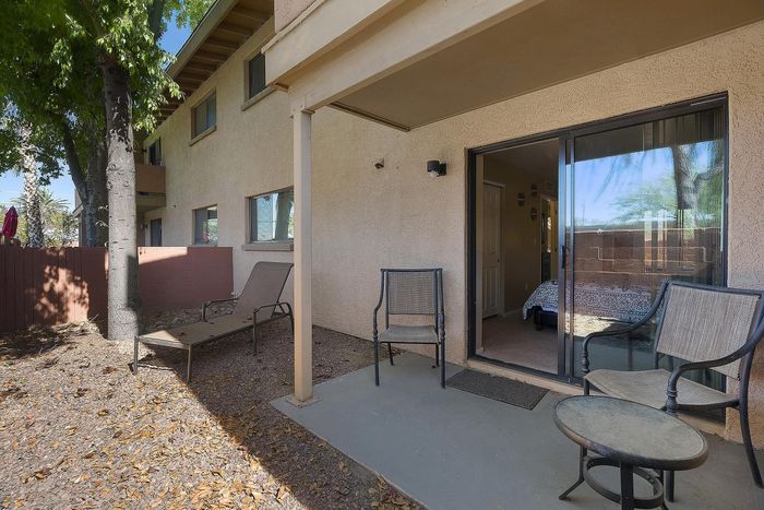 Charming apartment patio with cozy seating, lush trees, and a view into the inviting bedroom through sliding glass doors.