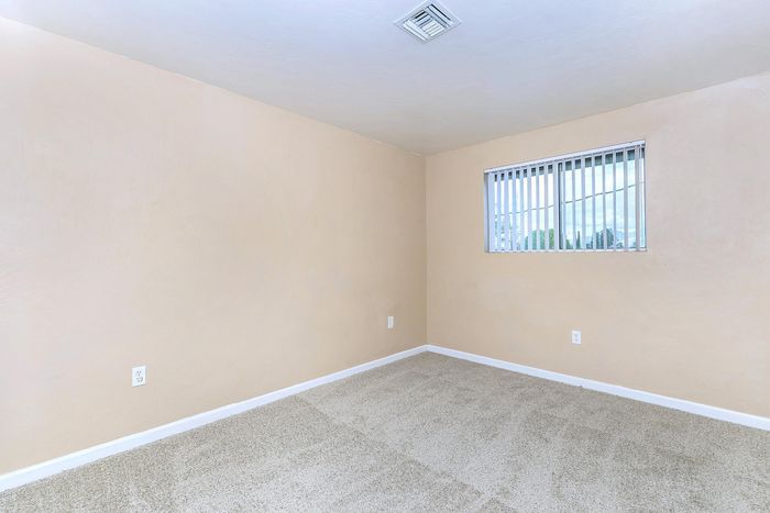 Bright and airy room with soft beige walls and plush carpet, featuring a large window with vertical blinds.