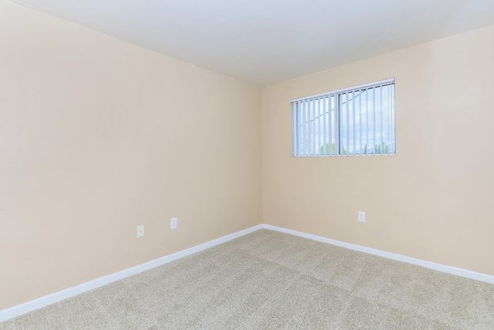 Bright and fresh empty room with beige walls and soft carpet flooring, featuring a large window with vertical blinds allowing natural light to fill the space.