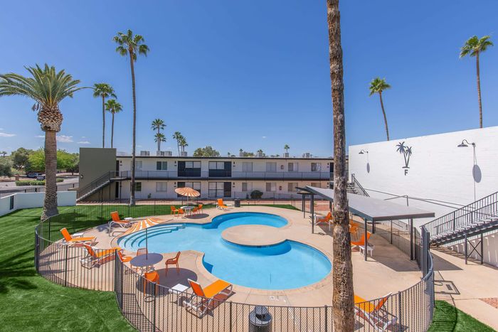 Inviting apartment complex featuring a sunlit swimming pool with stylish orange chairs and umbrellas, surrounded by lush palm trees and modern architecture.