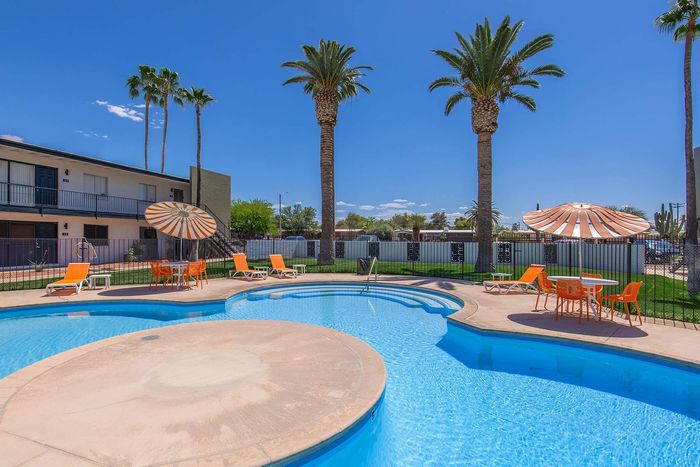 Inviting swimming pool area with bright orange lounge chairs, stylish umbrellas, and towering palm trees under a clear blue sky.