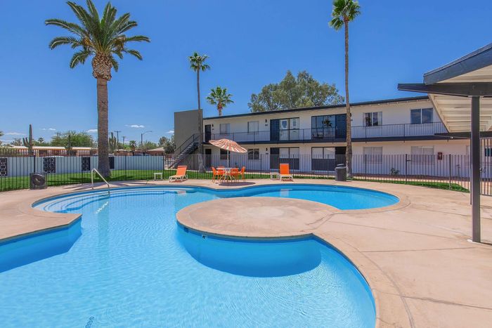 Inviting apartment complex with a stunning circular pool, vibrant orange seating, and towering palm trees under a clear blue sky.