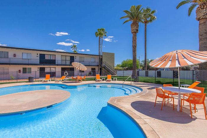 Inviting outdoor pool area with vibrant orange loungers and umbrellas surrounded by lush palm trees and a modern apartment complex.