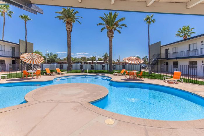 Inviting apartment complex with a sparkling pool, vibrant orange loungers, and charming umbrella-shaded seating.